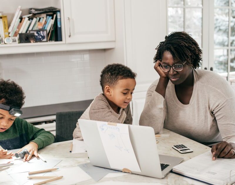 Parent working from home at a kitchen table with children, illustrating the challenge of separating work and home life.